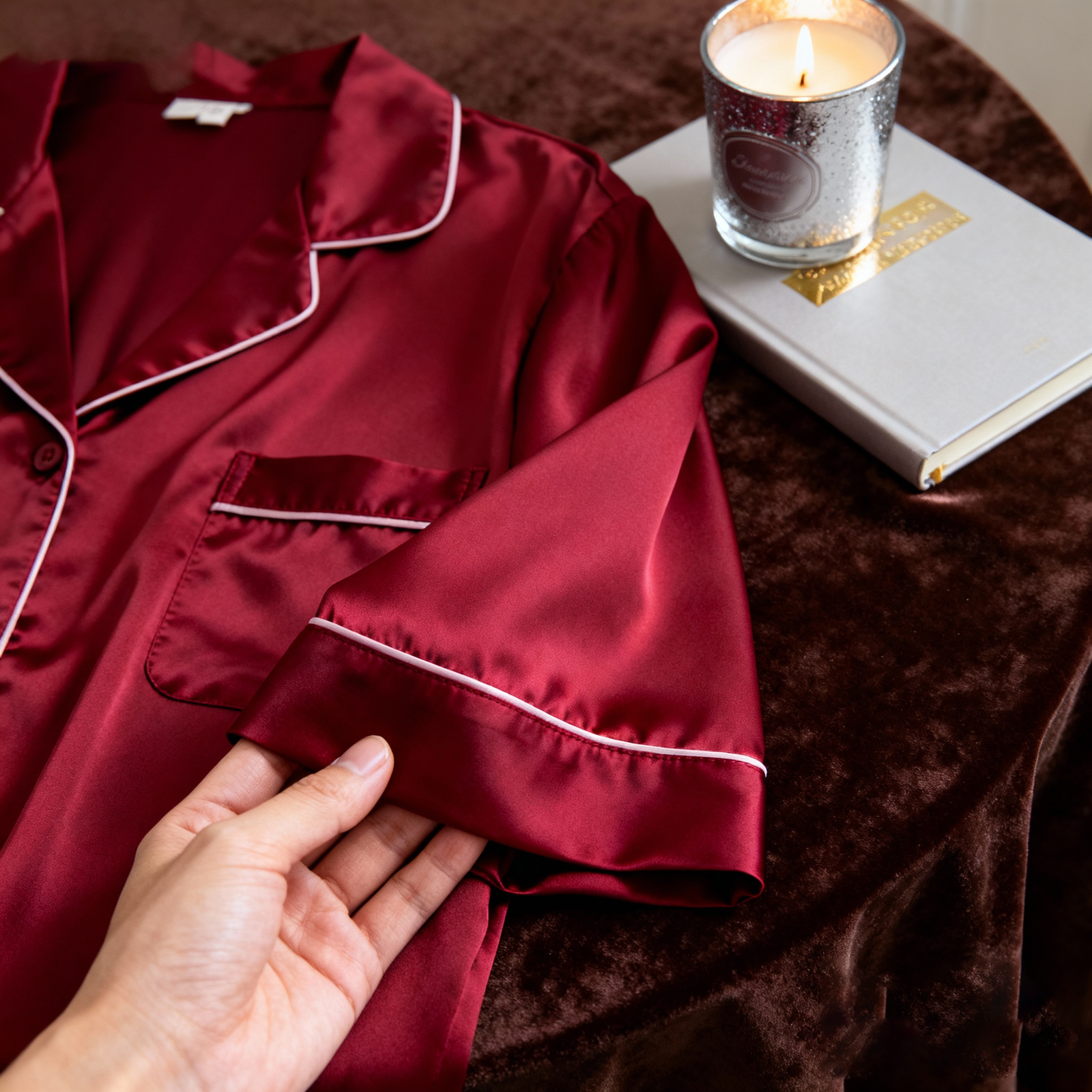 Burgundy silk pajama set with 细腻 sheen, fingers holding silk fabric corner on dark velvet background